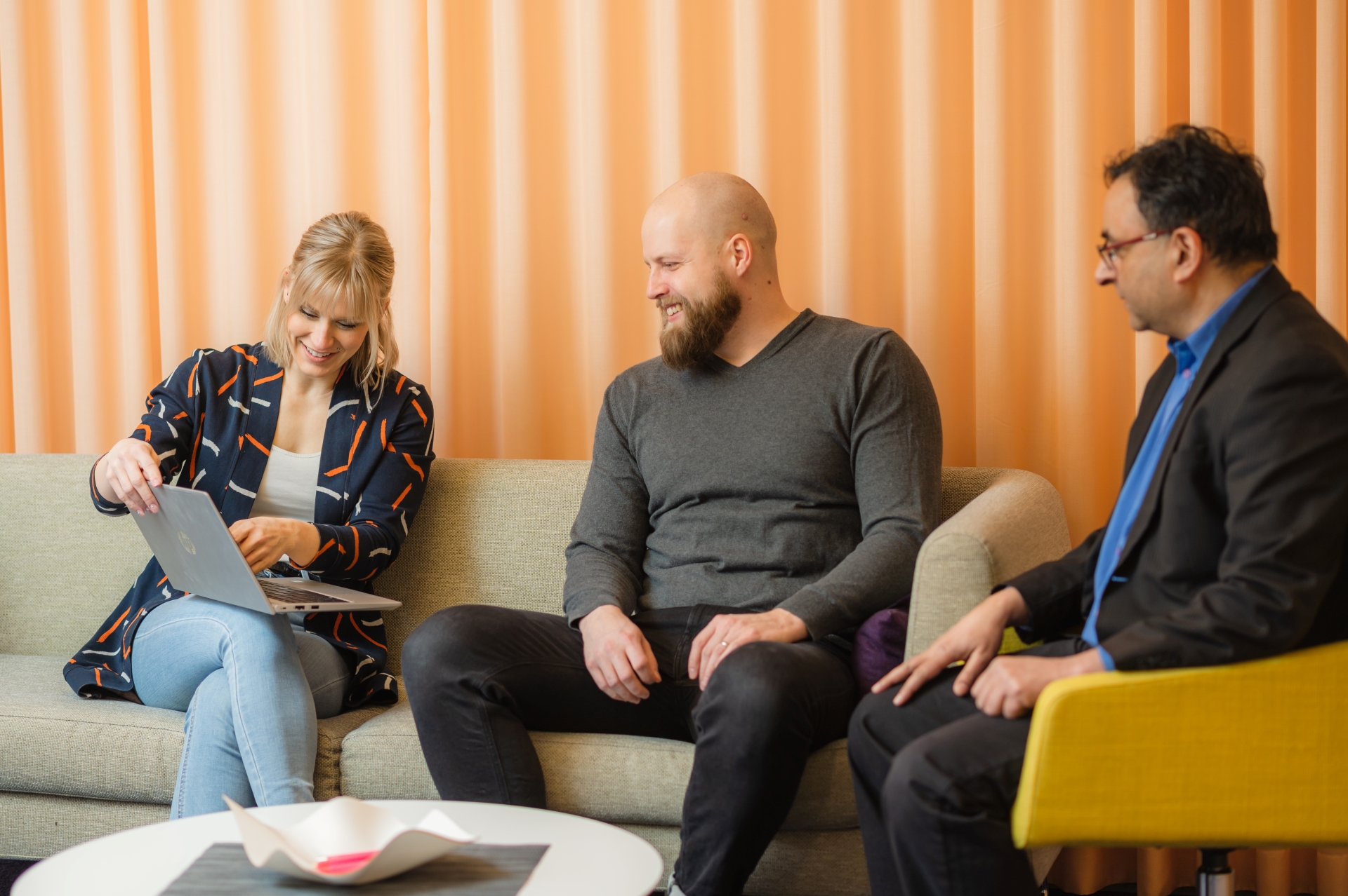 Kolme ihmistä istuu pöydän ääressä. Naisen esittelee kahdelle miehelle jotain kannettavalta tietokoneelta. Kaikki hymyilevät.