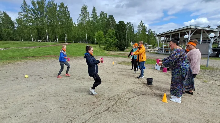 Aikuisia henkilöitä hiekkakentällä juosten ja seisoen pelaamassa peliä, jossa kuljetetaan jotain muovimukeissa.